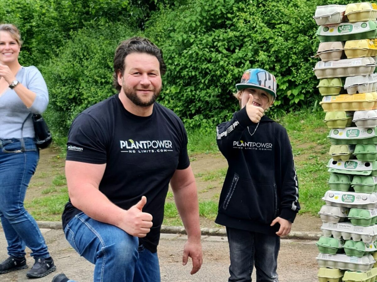 Das Foto zeigt Paul Schütte, den Gründer der Marke PlantPower-NoLimits, zusammen mit einem Kind. Beide tragen Fanartikel mit dem PlantPower-NoLimits-Logo, stehen vor einem Stapel Eierkartons und machen eine Daumen-hoch-Geste. Im Hintergrund sind grüne Büsche und eine Frau zu sehen.