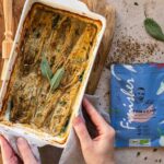 Eine weiße Auflaufform mit Chicorée in goldbrauner Senfsauce, dekoriert mit Kräutern und einem Salbeiblatt, neben einer Packung Bio Fisch & Scampi Food Finisher.