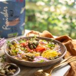 Nudelsalat mit gelber Paprika, roten Zwiebeln, Kapernäpfeln und Hähnchenstreifen in einer rustikalen Schale. Im Hintergrund ein natürlicher Garten und die Verpackung des Bio Fisch & Scampi Food Finishers.