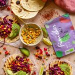 Mehrere gegrillte Tortilla-Wraps, belegt mit Avocado, Rotkohl, Bohnen und Mais, daneben eine Packung Bio Antipasti Food Finisher und Limettenspalten.