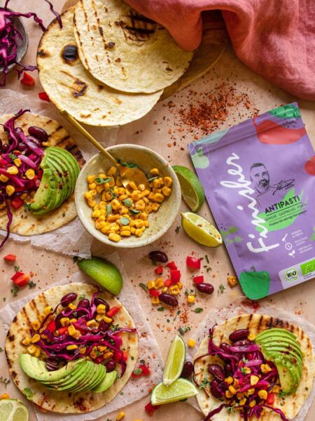 Mehrere gegrillte Tortilla-Wraps, belegt mit Avocado, Rotkohl, Bohnen und Mais, daneben eine Packung Bio Antipasti Food Finisher und Limettenspalten.