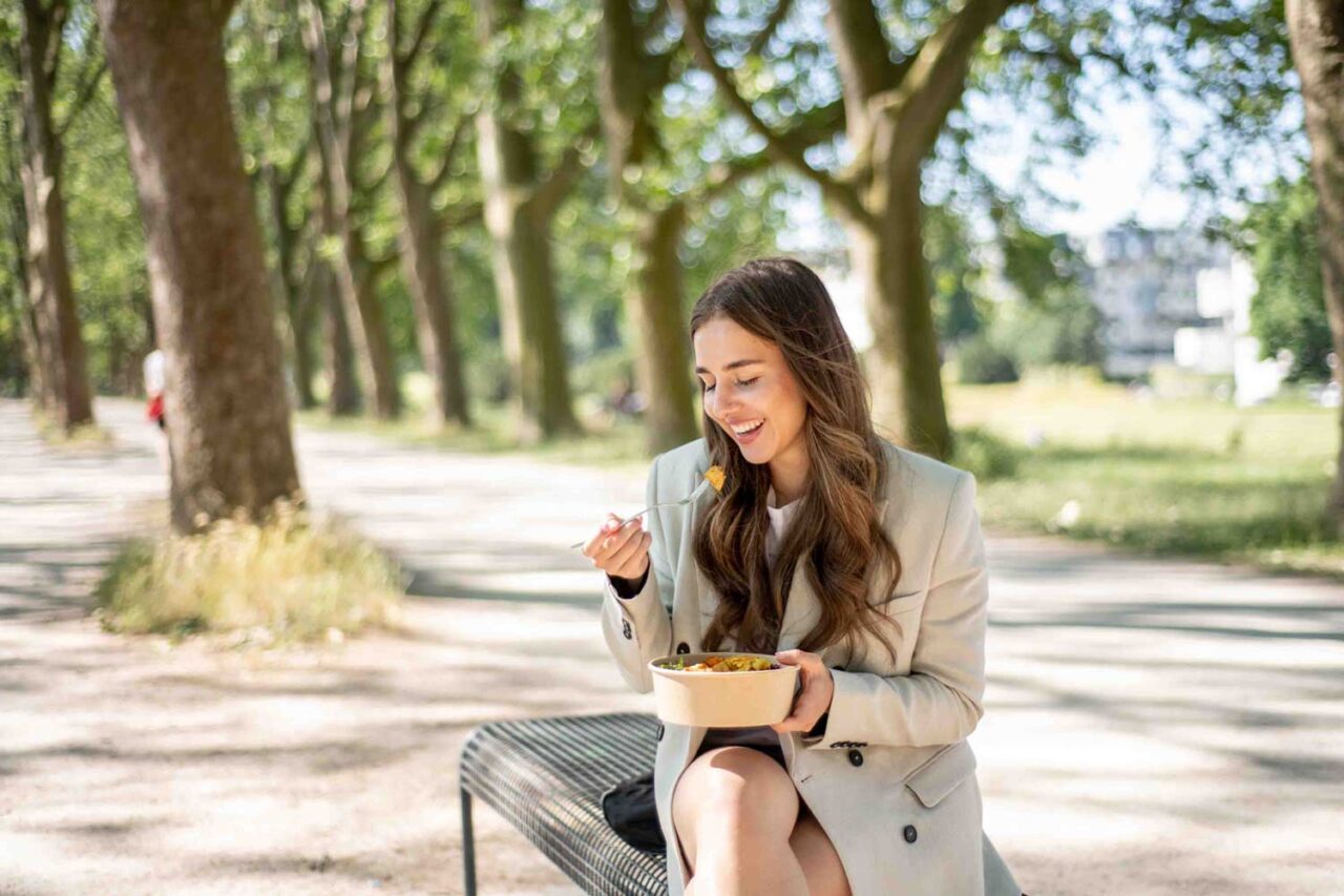 Eine junge Frau sitzt entspannt auf einer Parkbank und genießt einen gesunden, bunten Salat. Die frische Luft und die natürliche Umgebung unterstreichen ihren bewussten Ernährungsstil.