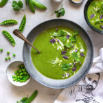Ein Teller mit grüner Erbsensuppe, dekoriert mit Sahne, frischen Kräutern und essbaren Blüten. Umgeben von frischen Erbsenschoten, Erbsen und Bio Braten-Magic Food Finisher in kleinen Schalen.
