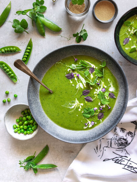 Ein Teller mit grüner Erbsensuppe, dekoriert mit Sahne, frischen Kräutern und essbaren Blüten. Umgeben von frischen Erbsenschoten, Erbsen und Bio Braten-Magic Food Finisher in kleinen Schalen.