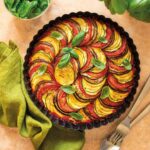 Eine aromatische Ratatouille mit Zucchini, Aubergine und Tomaten, dekorativ in einer Backform geschichtet und mit frischem Basilikum garniert, präsentiert mit grünem Tuch und Besteck. Ideal abgestimmt mit Bio Mediterran Food Finisher.