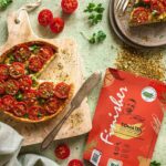 Fladenbrot mit Ricotta, Tomaten auf einem Holzbrett, daneben ein Teller mit einem Stück davon sowie die rote Verpackung des Bio Tomaten Food Finishers.