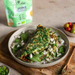 Ein goldbraunes Omelette Rezept mit Spinat und Feta auf einem Teller, daneben die grüne Verpackung des Bio Lecker-Ei Food Finishers mit der Illustration von Koch Luis Dias. Umgeben von frischen Zutaten und Gewürzen.