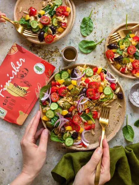 Drei Teller mit Orzo-Salat, bunt belegt mit Gemüse und gewürzt mit dem Bio Tomaten Food Finisher, dessen Verpackung gut sichtbar im Bild liegt.