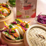 Sandwich Rezept. Frische Bao Buns gefüllt mit Pulled Jackfruit, Rotkohl, Karotten und Koriander, serviert in Bambusdämpfkörben. Perfekt abgestimmt mit Bio Espresso BBQ Food Finisher, der im Hintergrund zu sehen ist.