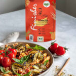 Ein Teller mit Tomaten-Aubergine-Pasta, dekoriert mit frischem Basilikum und Tomaten, daneben die rote Verpackung des Bio Tomaten Food Finishers.