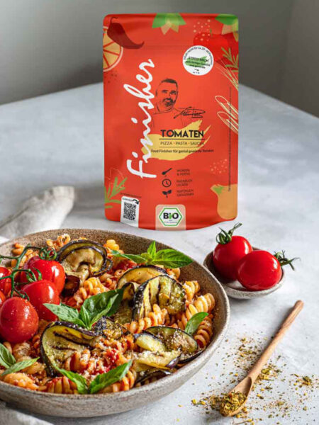 Ein Teller mit Tomaten-Aubergine-Pasta, dekoriert mit frischem Basilikum und Tomaten, daneben die rote Verpackung des Bio Tomaten Food Finishers.