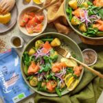 Ein Teller mit Räucherlachs, Kartoffeln, Zuckerschoten und roten Zwiebeln, daneben Zitronenschnitze, Gewürze und die blaue Verpackung des Bio Fisch & Scampi Food Finishers.