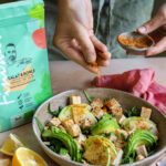 Bowl mit Tofu, Avocado, Zwiebeln, Kichererbsen und Rucola. Eine Hand streut den Bio Salat & Bowls Food Finisher darüber. Die Produktverpackung ist am Bildrand sichtbar.