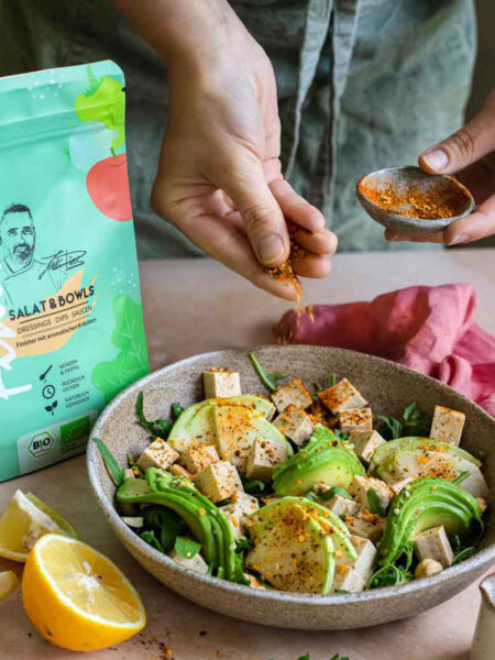 Bowl mit Tofu, Avocado, Zwiebeln, Kichererbsen und Rucola. Eine Hand streut den Bio Salat & Bowls Food Finisher darüber. Die Produktverpackung ist am Bildrand sichtbar.