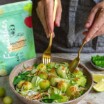 Rezept für Bowl mit Honigmelone, Reisnudeln, Babyspinat und Kräutern, mit dem Bio Salat & Bowls Food Finisher gewürzt. Zwei goldene Gabeln greifen in die Schale, die Produktverpackung ist sichtbar im Hintergrund