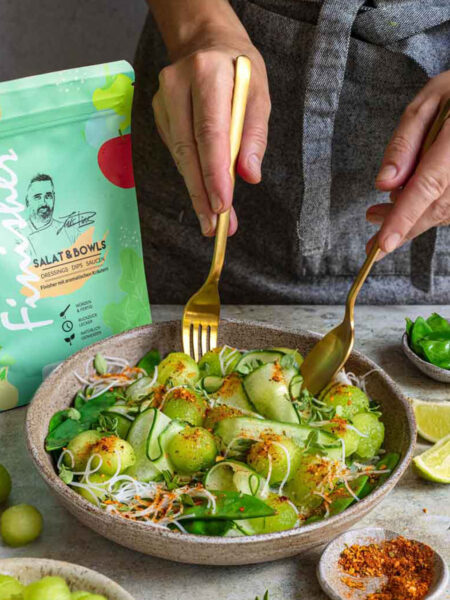 Rezept für Bowl mit Honigmelone, Reisnudeln, Babyspinat und Kräutern, mit dem Bio Salat & Bowls Food Finisher gewürzt. Zwei goldene Gabeln greifen in die Schale, die Produktverpackung ist sichtbar im Hintergrund