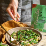 Eine Schale mit Tofu-Rührei, grünem Spargel und Erbsen, die mit Bio Lecker-Ei Food Finisher bestreut wird. Umgeben von frischem Gemüse und Kräutern, im Hintergrund die grüne Produktverpackung.