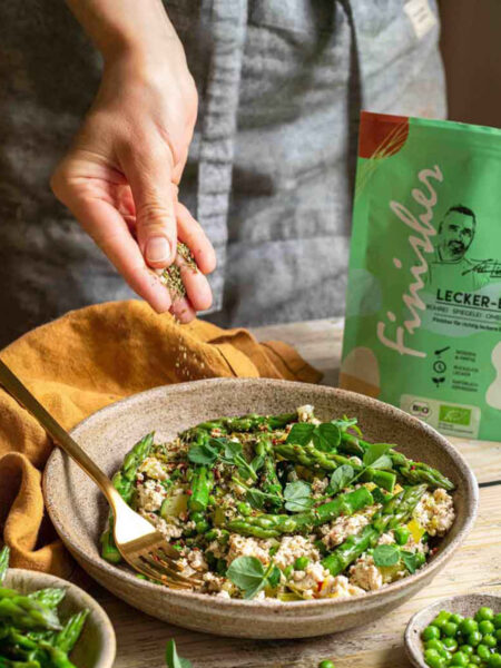 Eine Schale mit Tofu-Rührei, grünem Spargel und Erbsen, die mit Bio Lecker-Ei Food Finisher bestreut wird. Umgeben von frischem Gemüse und Kräutern, im Hintergrund die grüne Produktverpackung.