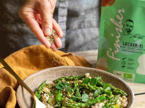 Eine Schale mit Tofu-Rührei, grünem Spargel und Erbsen, die mit Bio Lecker-Ei Food Finisher bestreut wird. Umgeben von frischem Gemüse und Kräutern, im Hintergrund die grüne Produktverpackung.