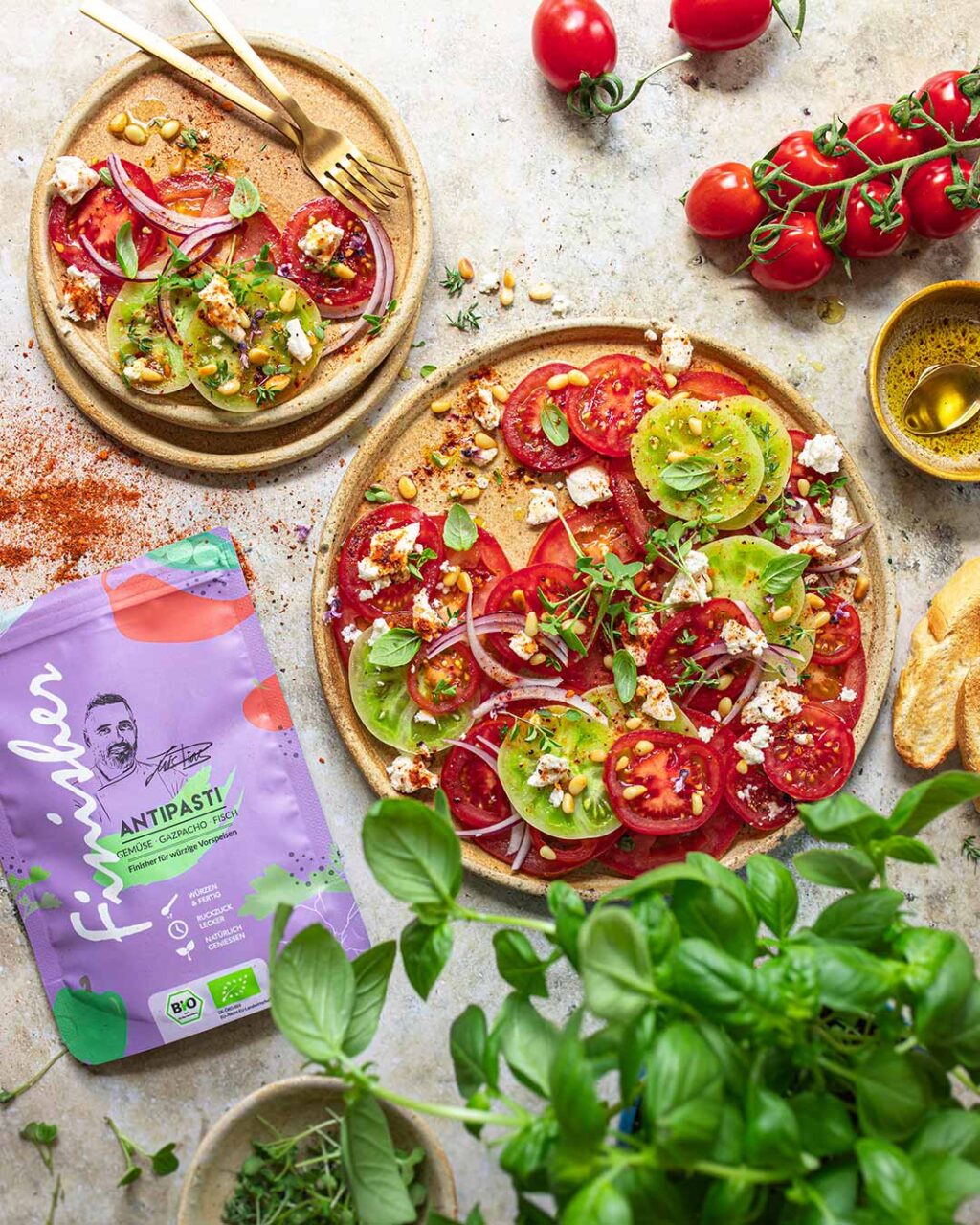 Rezemit für Ein großer Teller mit Tomaten Carpaccio aus bunten Tomatenscheiben, Feta und Zwiebeln, bestreut mit Bio Antipasti Food Finisher. Umgeben von frischem Basilikum, Tomaten und Brot.