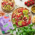 Rezemit für Ein großer Teller mit Tomaten Carpaccio aus bunten Tomatenscheiben, Feta und Zwiebeln, bestreut mit Bio Antipasti Food Finisher. Umgeben von frischem Basilikum, Tomaten und Brot.