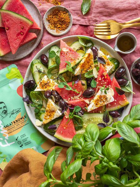 Rezept für Wassermelonen-Salat aus Melone, Feta, Oliven und Gurke, bestreut mit Bio Salat & Bowls Food Finisher. Daneben frische Zutaten und die grüne Produktverpackung.
