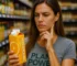 Junge Frau im grauen „Plant Power No Limits“ T-Shirt steht im Supermarkt vor Regalen voller Getränke und hält nachdenklich einen Saftkarton in der Hand.