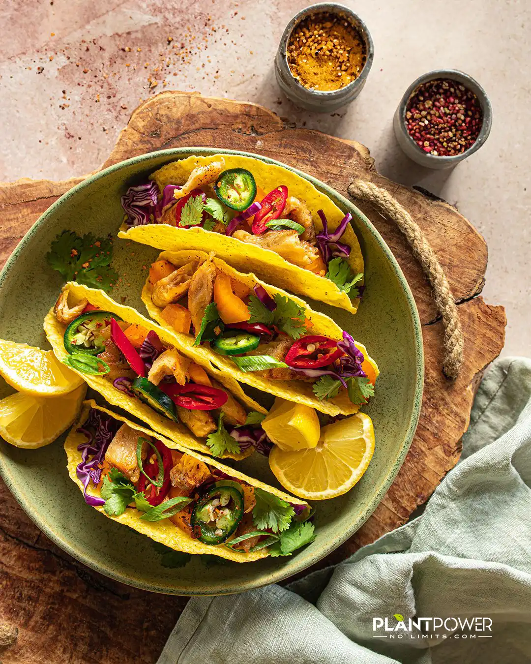 Veganer Tacos auf grünem Teller: gelbe Maistortillas gefüllt mit pflanzlichem Chicken, Rotkohl, Paprika, Jalapeños, Koriander und Zitronenvierteln, angerichtet auf rustikalem Holzbrett neben Gewürzdosen.
