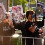 Aktivist*innen von Animal Equality demonstrieren hinter Absperrgittern; eine Person spricht durch ein Megafon, andere halten Plakate mit dem Porträt eines Konzernmanagers und dem Slogan „Chooses Profit Over Compassion“.