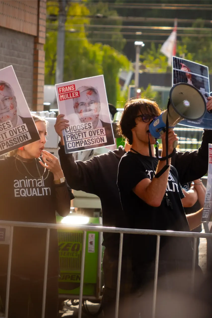 Aktivist*innen von Animal Equality demonstrieren hinter Absperrgittern; eine Person spricht durch ein Megafon, andere halten Plakate mit dem Porträt eines Konzernmanagers und dem Slogan „Chooses Profit Over Compassion“.
